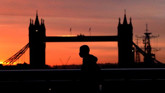 A person wearing a face mask walks across London Bridge, with Tower Bridge in the background.