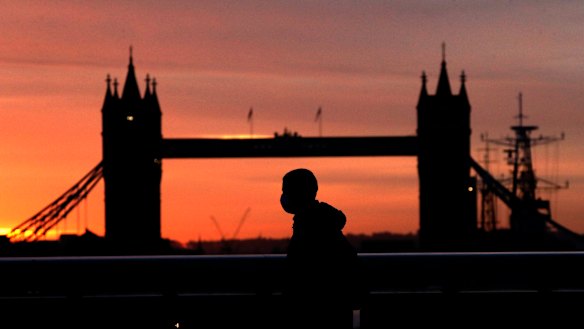 A person wearing a face mask walks across London Bridge, with Tower Bridge in the background.