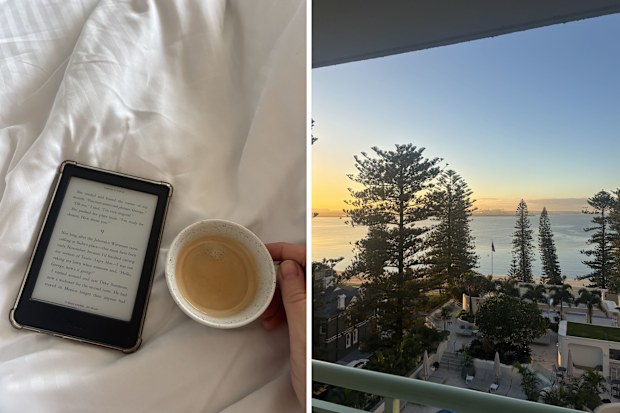 A hand holding a coffee on top of a bed next to an ereader and a photo of a view of the ocean from a hotel room