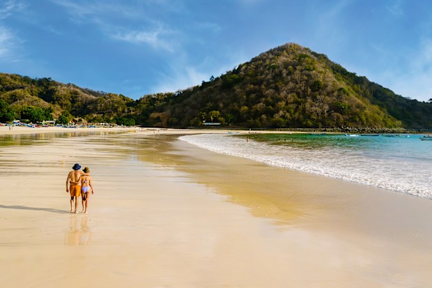 Young tourist couple walking together along tropical beach in Indonesia. Romantic walk by the shoreline with ocean waves and distant hills. Love and travel concept.