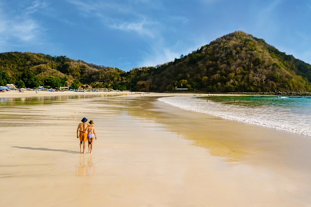 Young tourist couple walking together along tropical beach in Indonesia. Romantic walk by the shoreline with ocean waves and distant hills. Love and travel concept.