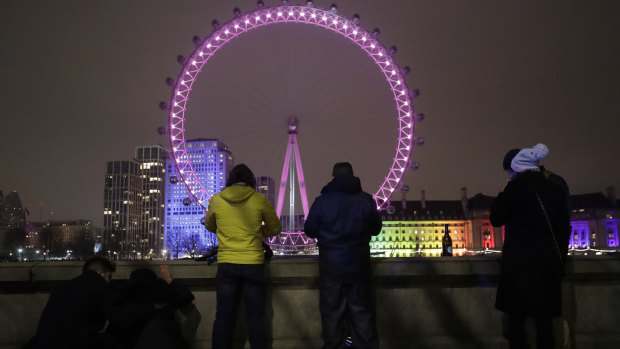 A small group of people look across from the embankment towards the London Eye Ferris wheel by the River Thames.