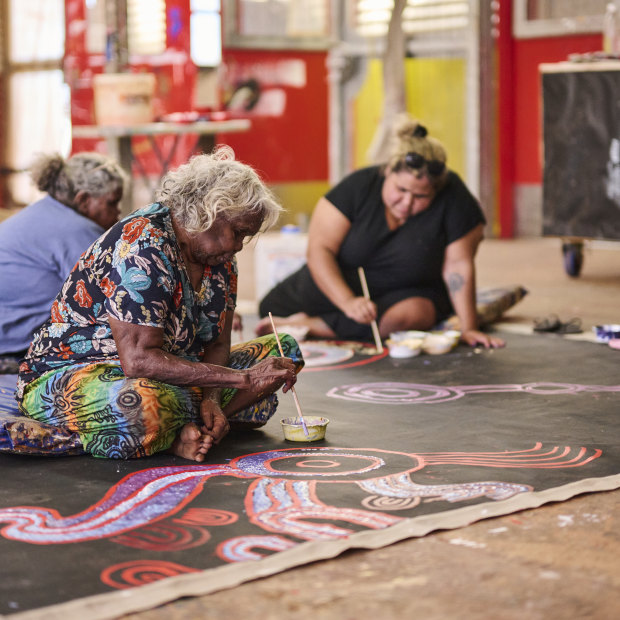 On the canvas, from left: Tuppy Goodwin, Puna Yanima, Sally Scales and Iluwanti Ken collaborate at Mimili Maku Arts.
Yaritji Young’s husband Frank sits on the chair.