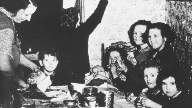 A woman slices bread for the eight children crowded around a table at meal-time. 