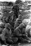 Argentinian soldiers captured at Goose Green, The Falklands Islands, being guarded by a British Royal Marine as they await transit out of the area.