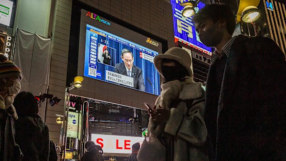 People walk through the Shinjuku area as the Prime Minister is seen on a large screen announcing the state of emergency.