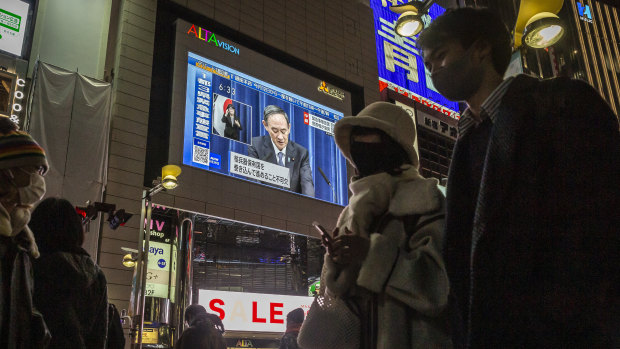 People walk through the Shinjuku area as the Prime Minister is seen on a large screen announcing the state of emergency.