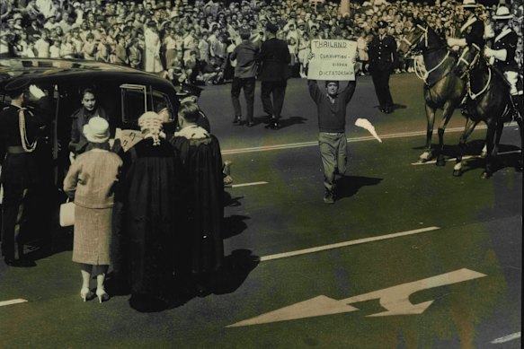 Enquanto a Rainha Sirikit desce do carro ao chegar a Sydney, em agosto de 1962, um manifestante atravessa a George Street com um cartaz, enquanto um segundo homem é conduzido para fora da estrada por um policial ao fundo.