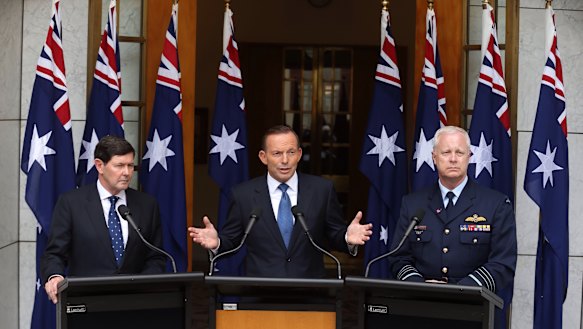 Then-prime minister Tony Abbott in 2015 announcing preparation for a troop deployment to Iraq. He is joined by then-Defence Minister Kevin Andrews and former Defence Force chief Air Chief Marshal Mark Binskin, and a number of national flags.