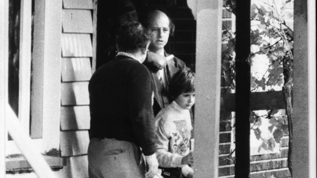 Senior Detective Peter Gill is held with a knife at his throat in the house doorway as the freed boy joins his father (back to camera)