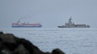 A UAE navy ship sails next to a cargo ship in the Strait of Hormuz. The key waterway remains effectively closed.