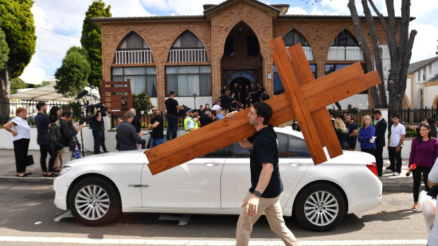 A man carrying crosses walks past Prime Minister Scott Morrison's car after Good Friday Easter services at St Charbel's Catholic Maronite Church at Punchbowl. 