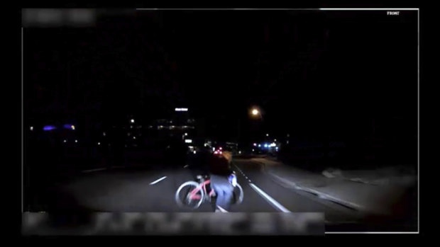 A camera inside the car shows a woman pushing a bicycle across the road in the dark. 