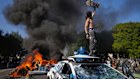 A protester stands on a burning taxi in LA.