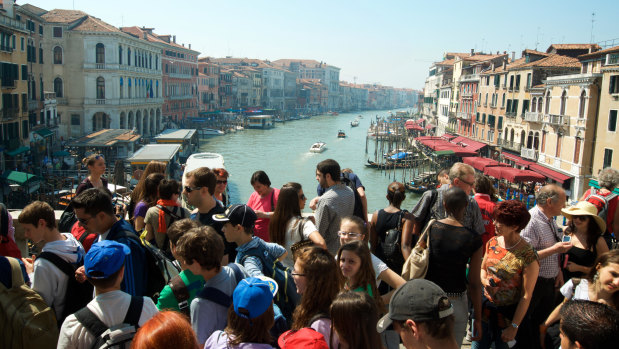 Crowds of tourists pass along the Rialto Bridge against a view of the Grand Canal in Venice.