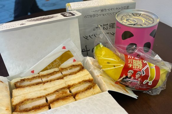 Tokyo vending machine haul: a pork katsu sandwich, tinned strawberry cake, a banana and a mystery box containing biscuits and a typed message in Japanese. 