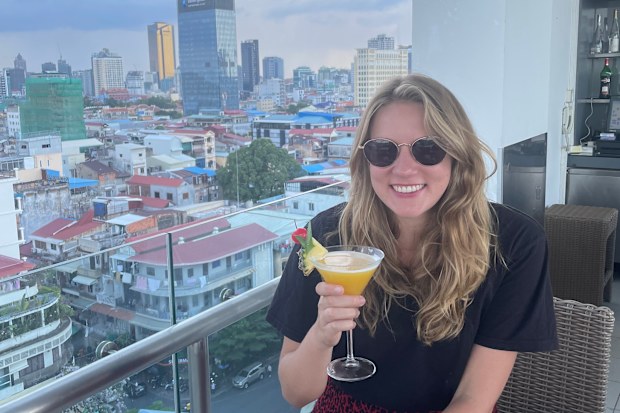 A woman sitting by the edge of a rooftop with views of a city, holding a cocktail 