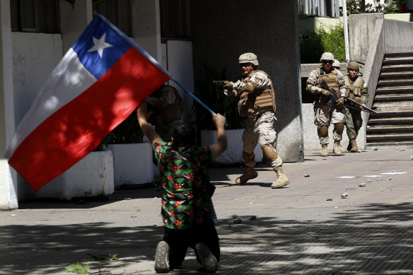 A demonstrator on his knees waves a Chilean flag as riot police officers clash with demonstrators.