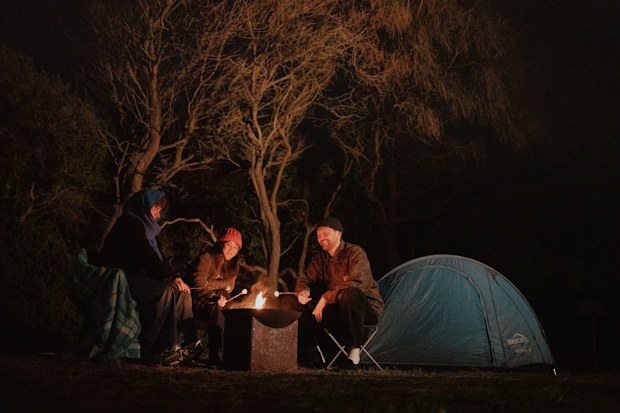 Three people sit around a campfire at night toasting marshmallows with a tent beside them. 