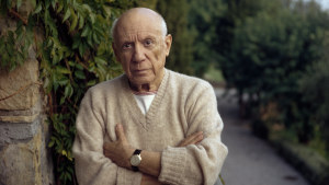 1966 : Pablo Picasso standing by a green fern with folded arms, wearing a cashmere sweater. 