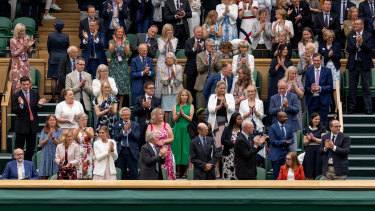 Członkowie rodziny królewskiej i widzowie stoją obok Sarah Gilbert, siedzącej w prawym dolnym rogu na stadionie Wimbledon Central. 