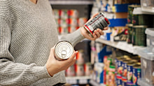 Stock image of a woman holding tins in the supermarket aisle.