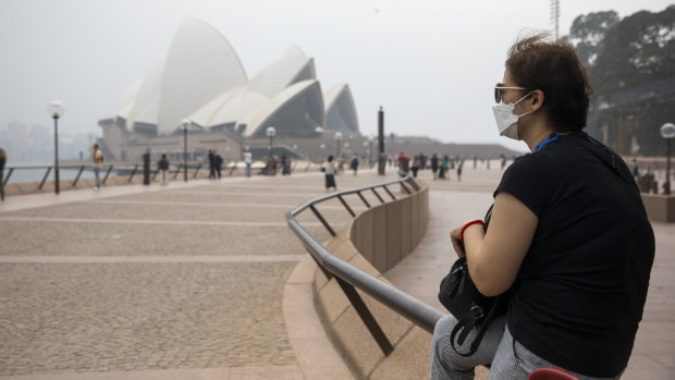 A woman wearing a face mask sits near a haze-shrouded Sydney Opera House.
