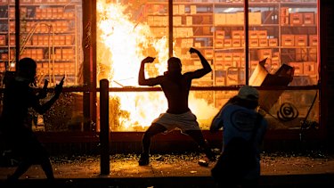 A man poses for photos in front of a fire at an AutoZone store, while protesters hold a rally for George Floyd in Minneapolis on Wednesday.