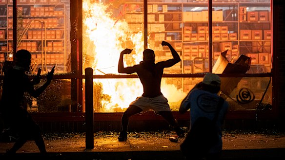 A man poses for photos in front of a fire at an AutoZone store, while protesters hold a rally for George Floyd in Minneapolis on Wednesday.