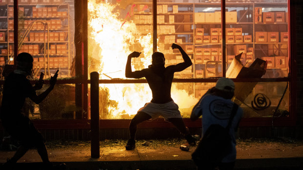 A man poses for photos in front of a fire at an AutoZone store, while protesters hold a rally for George Floyd in Minneapolis on Wednesday.