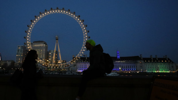 People chat as the London Eye is illuminated in yellow to signify a “beacon of remembrance” in London, one year since PM Johnson announced a national lockdown.