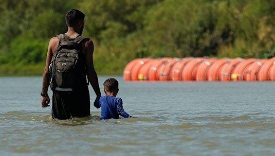 Migrants walk past large buoys being used as a floating border barrier on the Rio Grande River between Texas and Mexico.