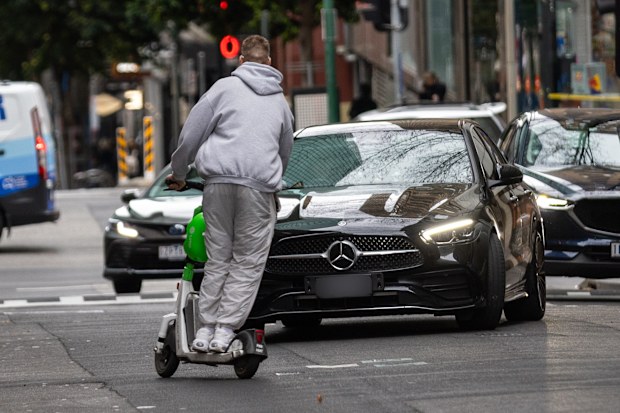 A man rides an E scooter the wrong way down Little Bourke with no helmet, swerving to miss an oncoming car.