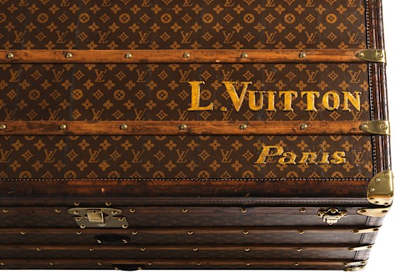 Uma visão detalhada de um baú de lona feminino com monograma de 1910, mostrando a personalização e as ferragens originais pintadas à mão que inspiraram a herança de viagens da Maison.