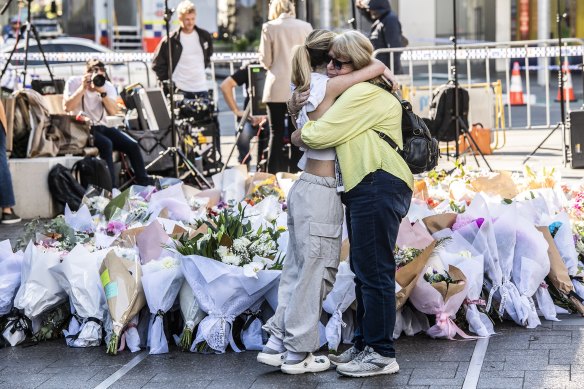 People embrace in front of the growing stack of floral tributes at the site of the Bondi Junction attacks.