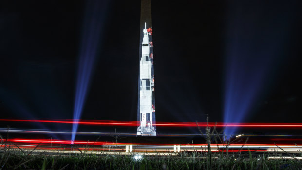 The lights of passing cars streak the foreground as the image of a Saturn V rocket is projected on the east face of the Washington Monument.