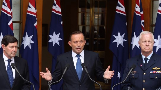 Then-prime minister Tony Abbott in 2015 announcing preparation for a troop deployment to Iraq. He is joined by then-Defence Minister Kevin Andrews and former Defence Force chief Air Chief Marshal Mark Binskin, and a number of national flags.
