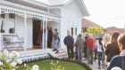 A line of people wait to be let into a white weatherboard property with a white picket fence and manicured garden.