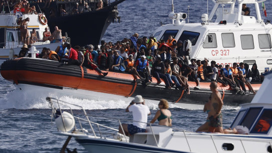 An Italian Coast Guard boat carries migrants as tourists on a boat, foreground, watch, near the port of the Sicilian island of Lampedusa, in southern Italy.