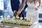 Newlyweds happily cut, laugh and taste the wedding cake. Beautiful Bride and Groom Celebrate Wedding at an Evening Reception Party with Multiethnic Friends. Married Couple Standing at a Dinner Table, Cutting Wedding Cake.