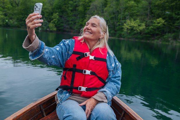 A smiling senior woman in a denim jacket and life vest takes a selfie with her smartphone while enjoying a boat ride on a tranquil green lake.