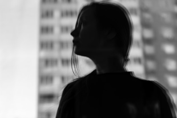 Moody black and white portrait of a young woman in silhouette, contemplating a modern city landscape through a window. Represents urban isolation, mental well-being. Minimal cinematic female portrait.