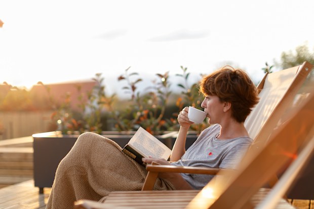 woman is sitting on patio on the deck chair and enjoying surrounding while drinking coffee or tea and reading a book