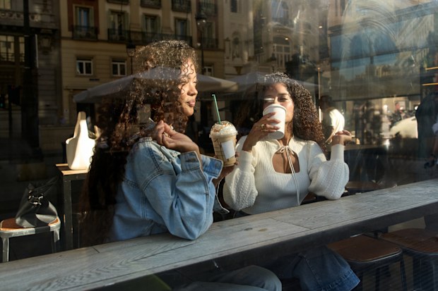 Two adult women sharing drinks and conversation inside a urban cafe, seen through the window with city reflections