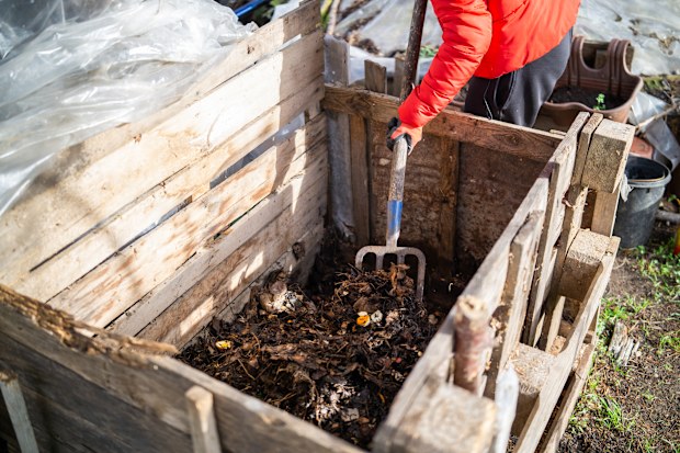 A person wearing gloves uses a garden fork to turn and aerate decomposing organic material inside a wooden compost bin.