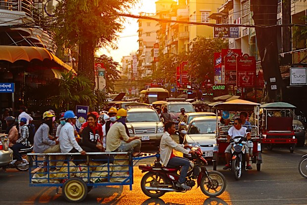 A busy city street at sunset with a man on a motorbike towing a trailer full of people, tuk tuks, and cars
