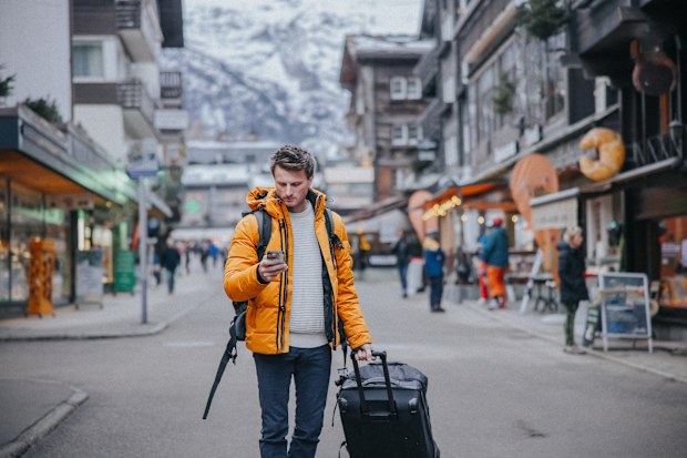 Man traveler using mobile phone while pulling luggage trolley on street to search an address of his stay in a town at Switzerland