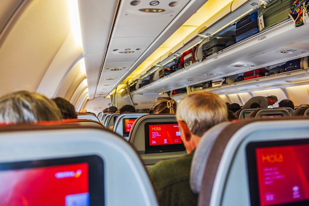 Close up view of passengers sitting inside Iberia Airlines cabin with seatback screens and luggage in overhead compartments.