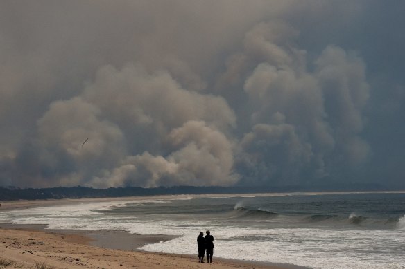 People stand on Diamond Beach watching smoke from bushfires in the Wallabi Point area fill the sky.