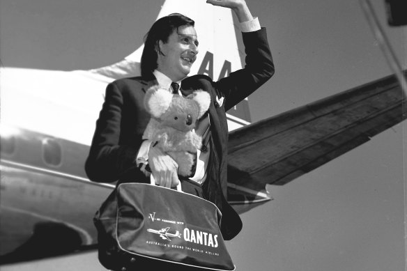 Barry Humphries holding a koala toy and Qantas bag waving on arrival in Melbourne.